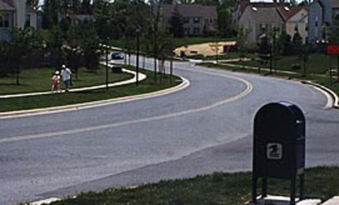 photo shows a residential street with two wide lanes.  The street slopes gently down to the right for about a half a block, then turns left and goes uphill -- we can see the street for about a half block beyond the bend at the bottom of the hill.